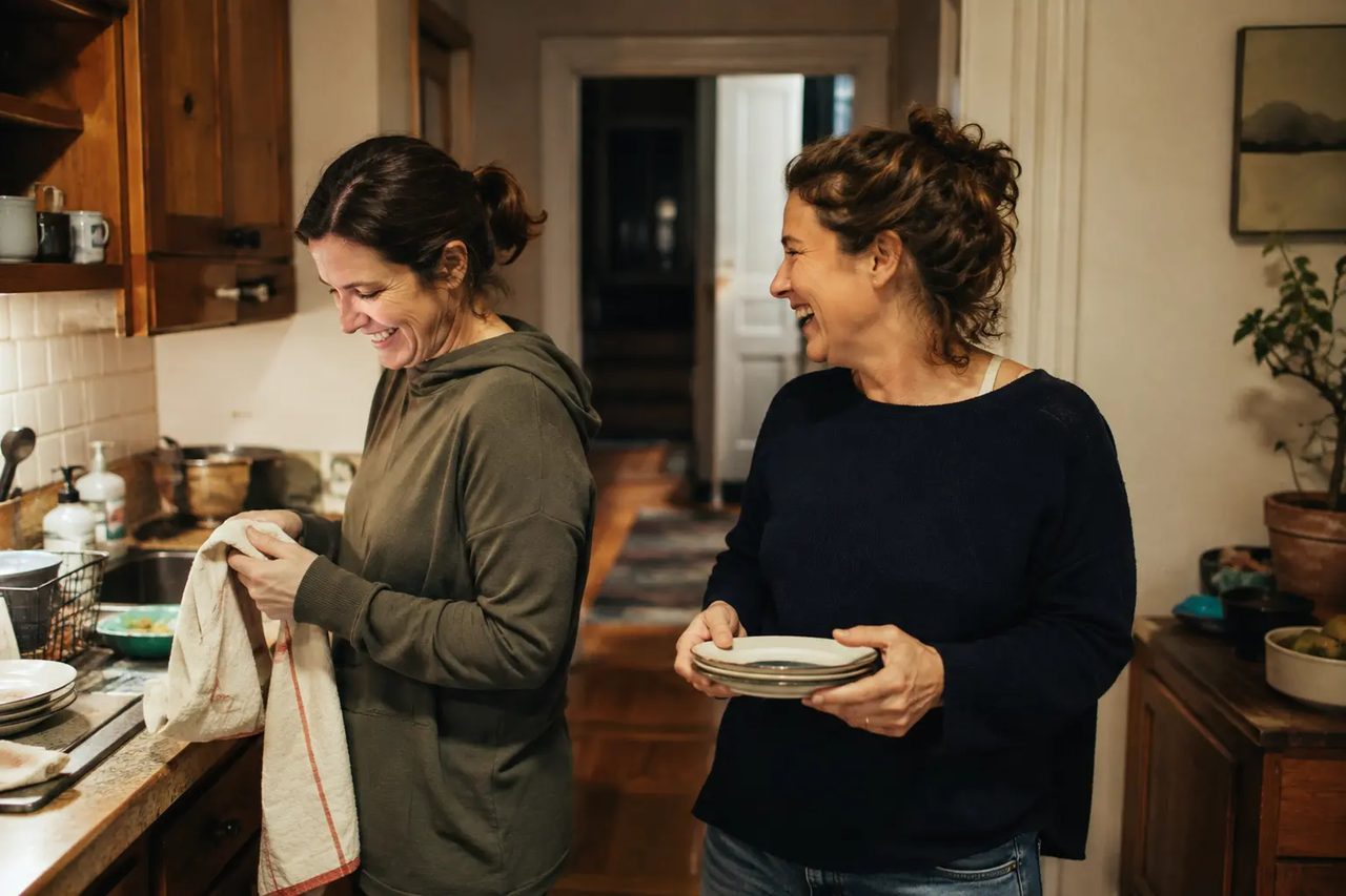 Two friends laughing together while putting the dishes away in a kitchen.