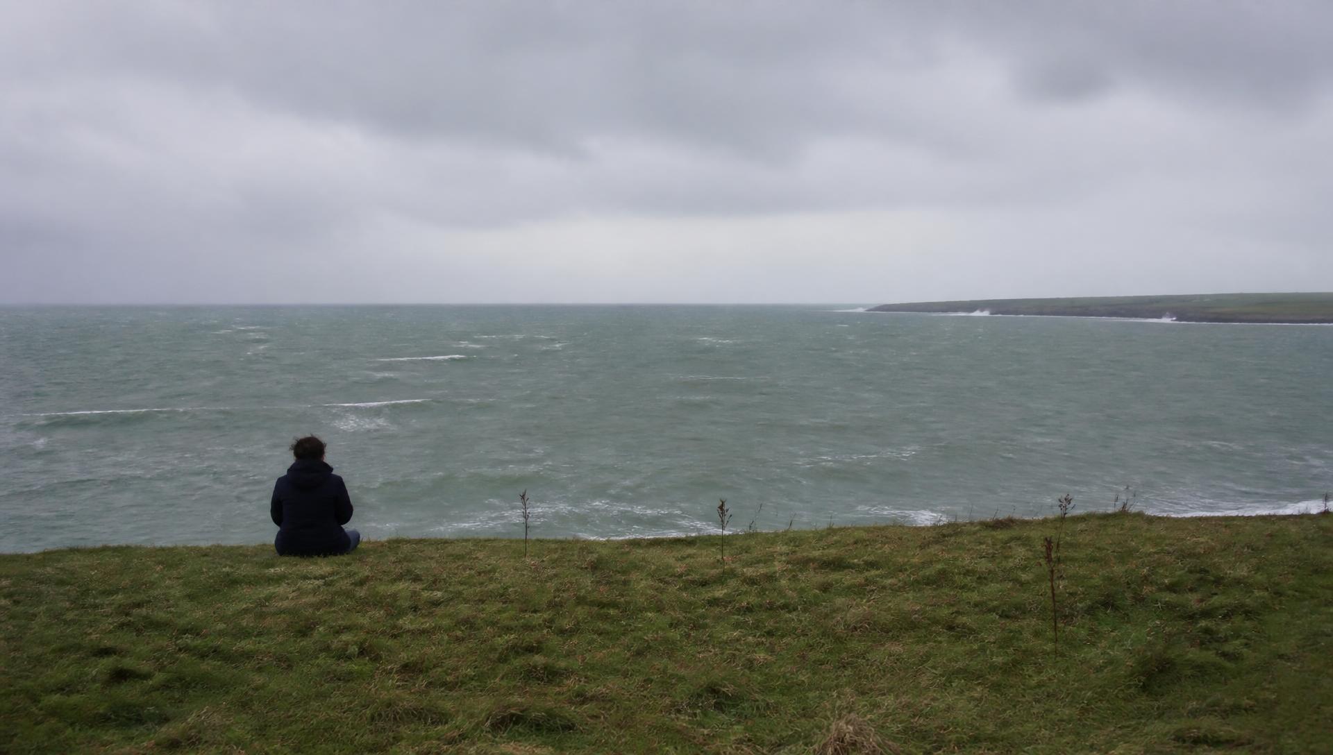 A person sitting alone on grass above a grey sea, looking out at the horizon