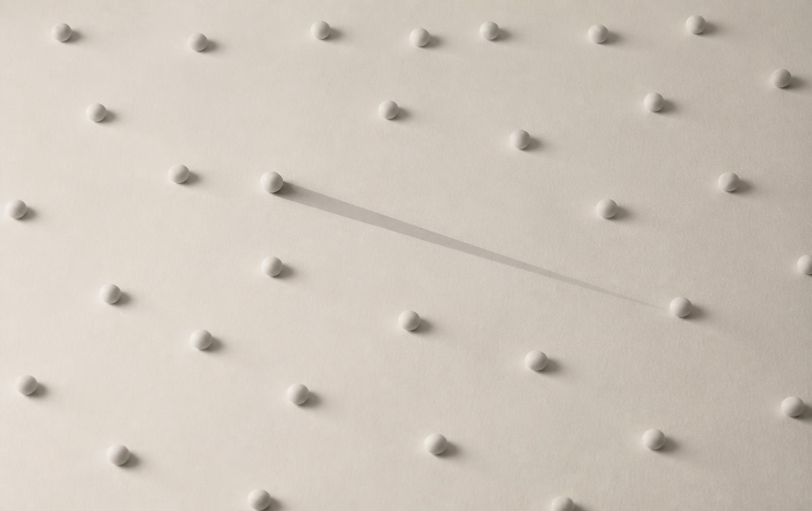 Dozens of identical pale pebbles on a matte surface, one casting a long shadow the others do not