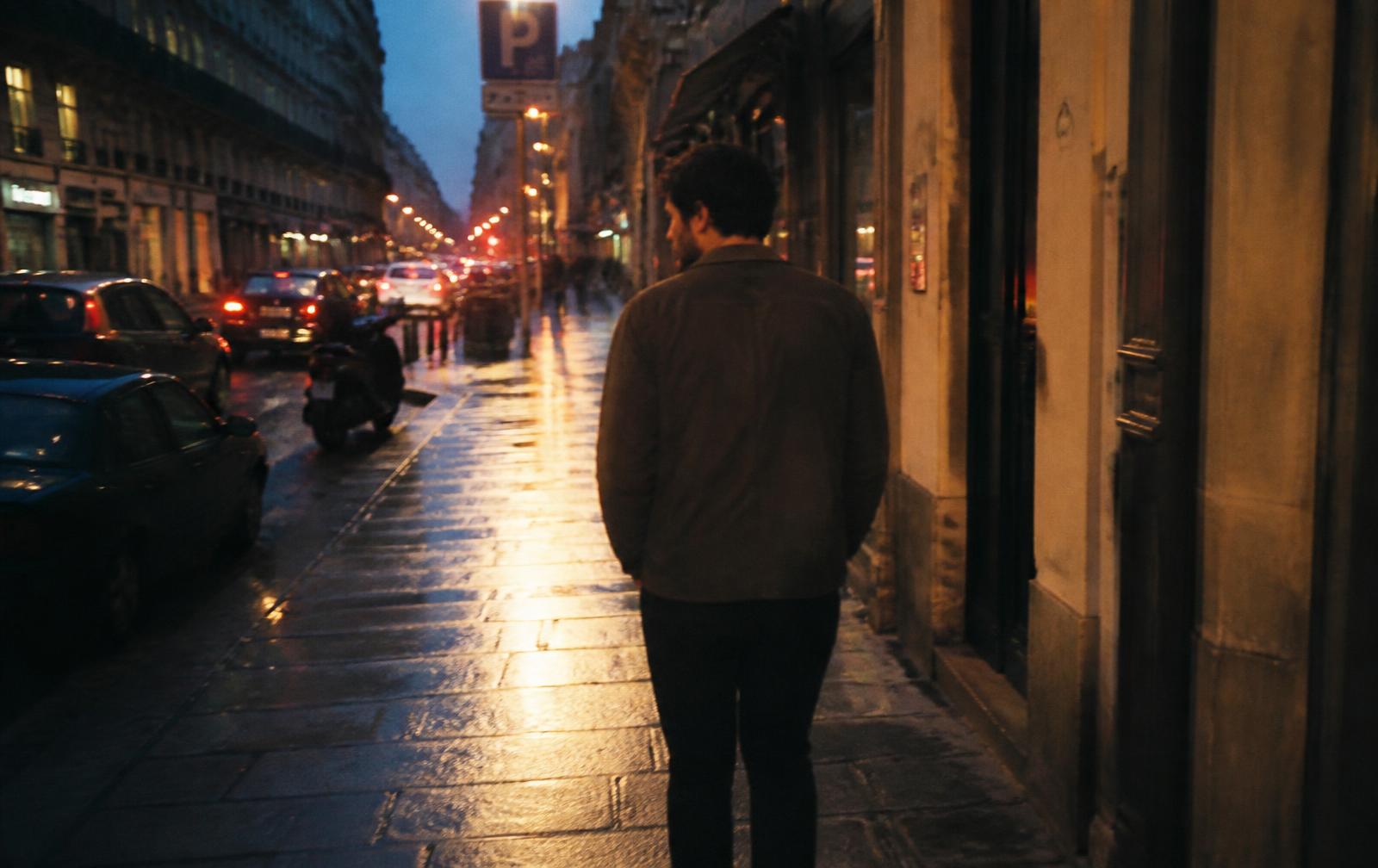 A man in his early thirties walking alone through a city street at dusk, seen from behind